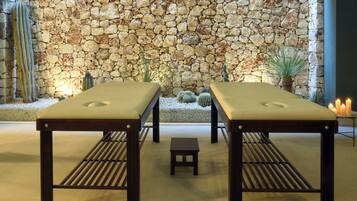 Two wooden massage tables with cushions, a small stool, and a stone wall with cacti in the background.