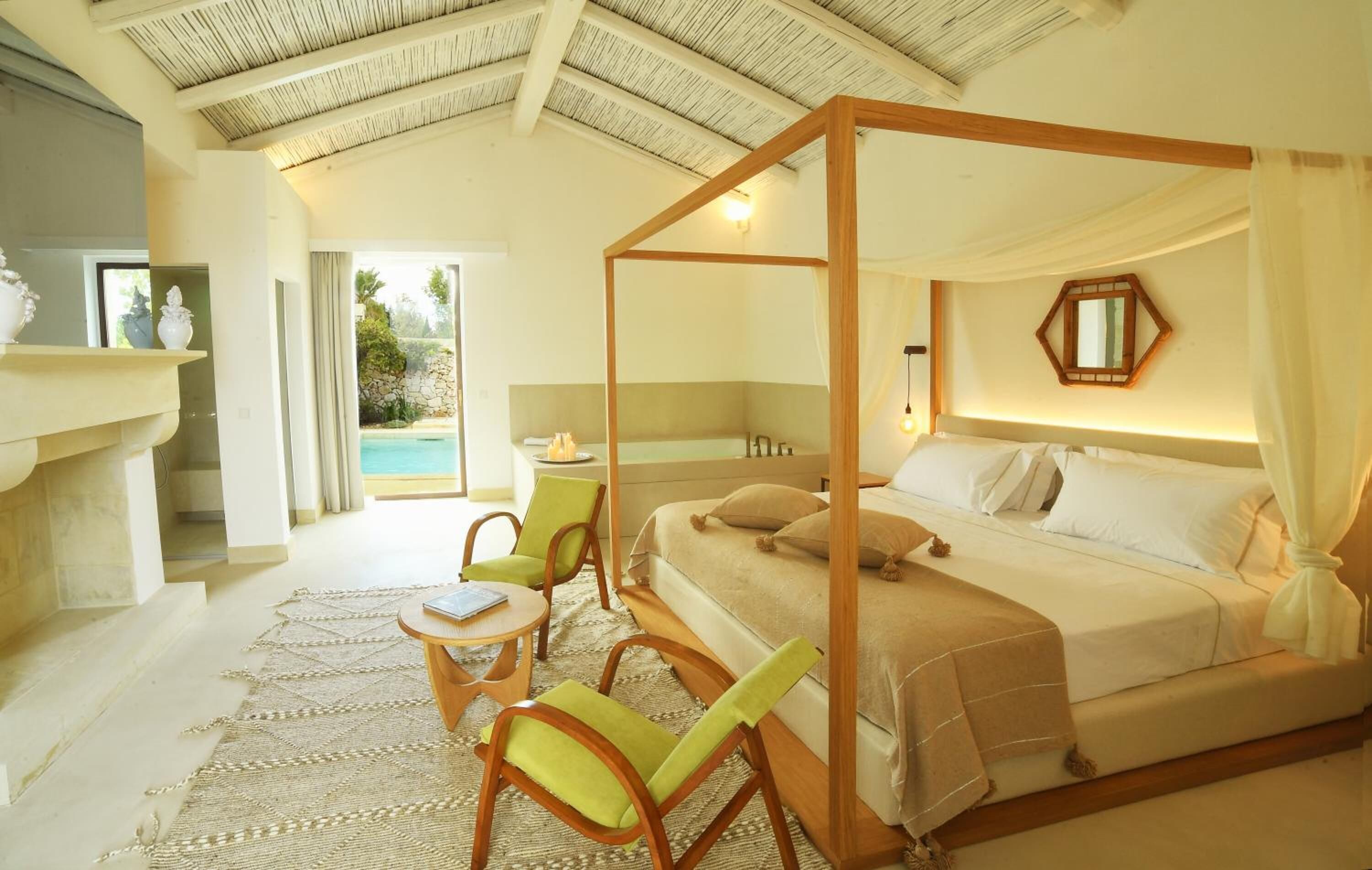 A spacious bedroom with a four-poster bed, a sitting area with chairs, and a view of a swimming pool.