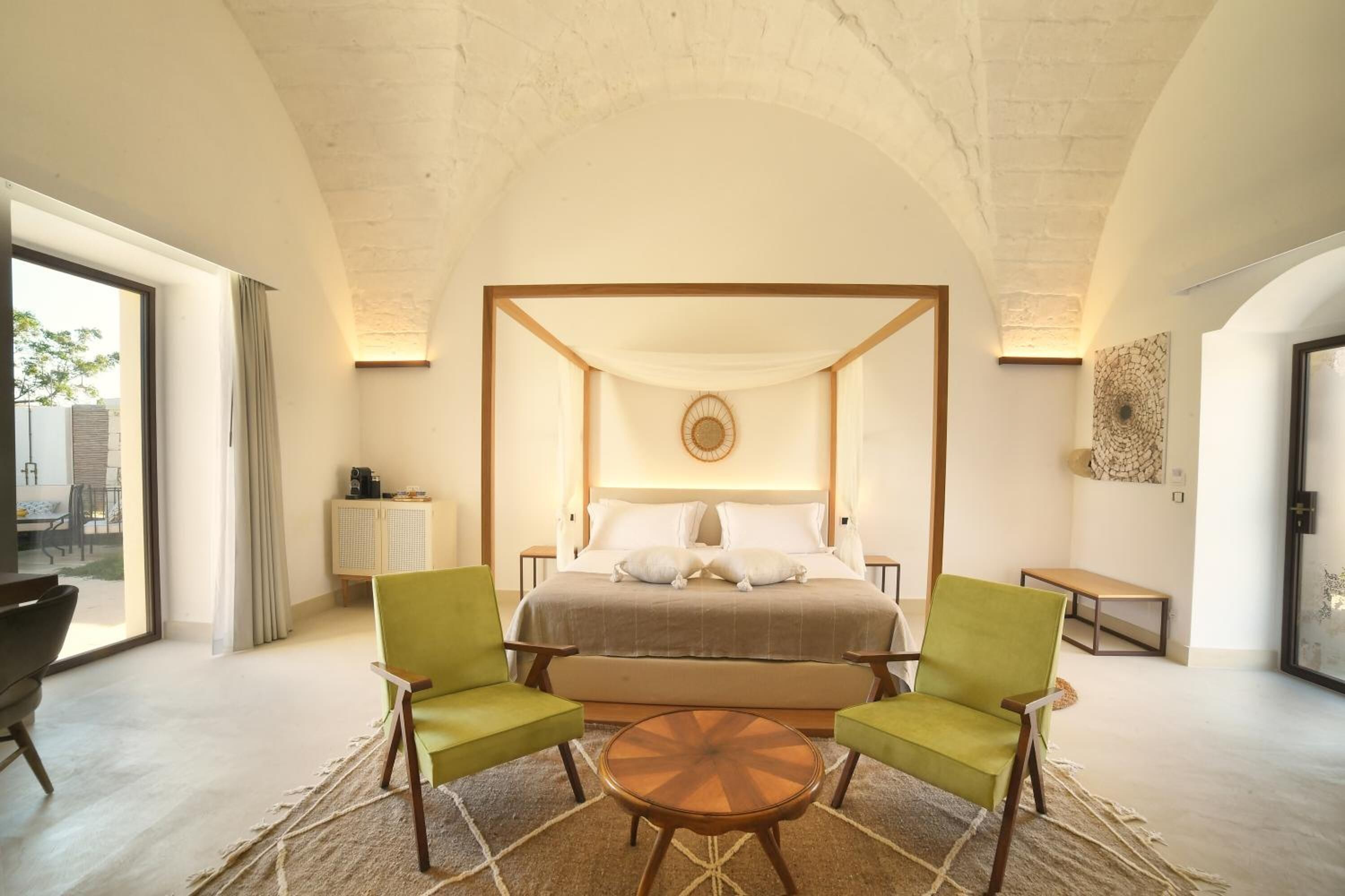 A bedroom with a four-poster bed, two green chairs, a round coffee table, and a view of the outdoors.