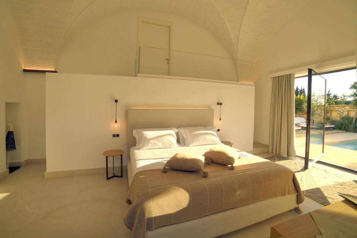 Een slaapkamer met een groot bed, een klein nachtkastje, een nachttafel en uitzicht op een zwembad door een glazen deur.