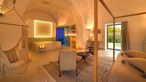 A cozy, well-lit room with a hammock, a sofa, a small table, and a view of the outdoors through a large window.