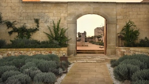 An arched entrance to a courtyard with stone walls, surrounded by neatly trimmed shrubs and a stone pathway.