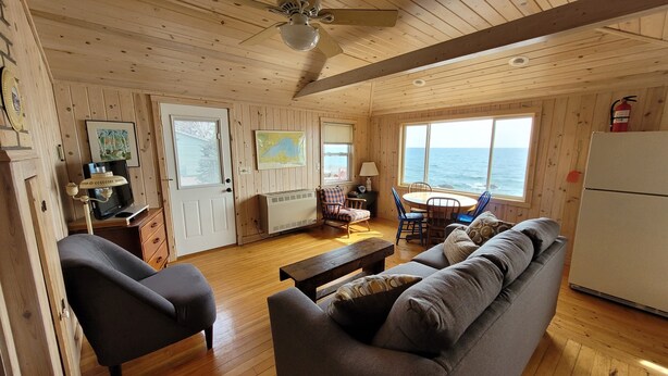 Una cabaña acogedora con paredes y suelos de madera, un comedor con vistas al océano y una sala de estar con sofá y sillones.