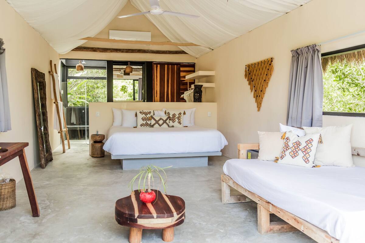 Una habitación con cama, sofá, un taburete de madera, ventilador de techo y ventana con cortinas.