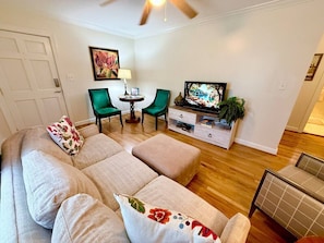 A living room with a beige sectional sofa, two green chairs, a small table, a television, and a plant. - Chic Condo in Downtown Raleigh (Raleigh)