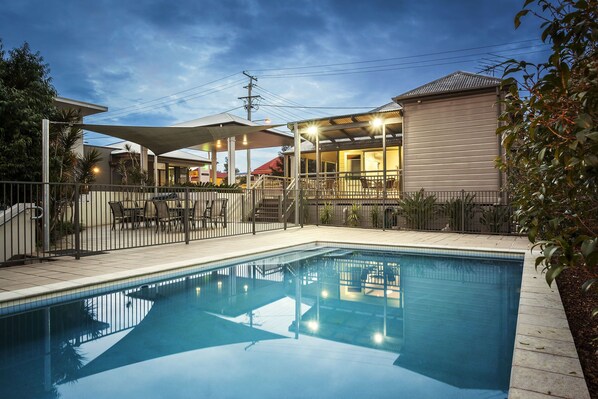 A swimming pool with a covered patio area, a fenced deck, and a building with a sloped roof. - Quest Ipswich (Ipswich)