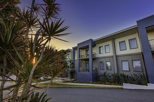 Un edificio moderno de varios pisos con balcones, techo plano y un jardín frontal ajardinado con palmeras.