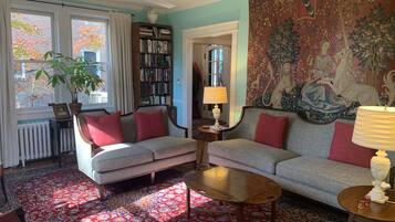 A living room with a sofa, armchair, coffee table, and a bookshelf. There is a large tapestry on the wall and a window with curtains.