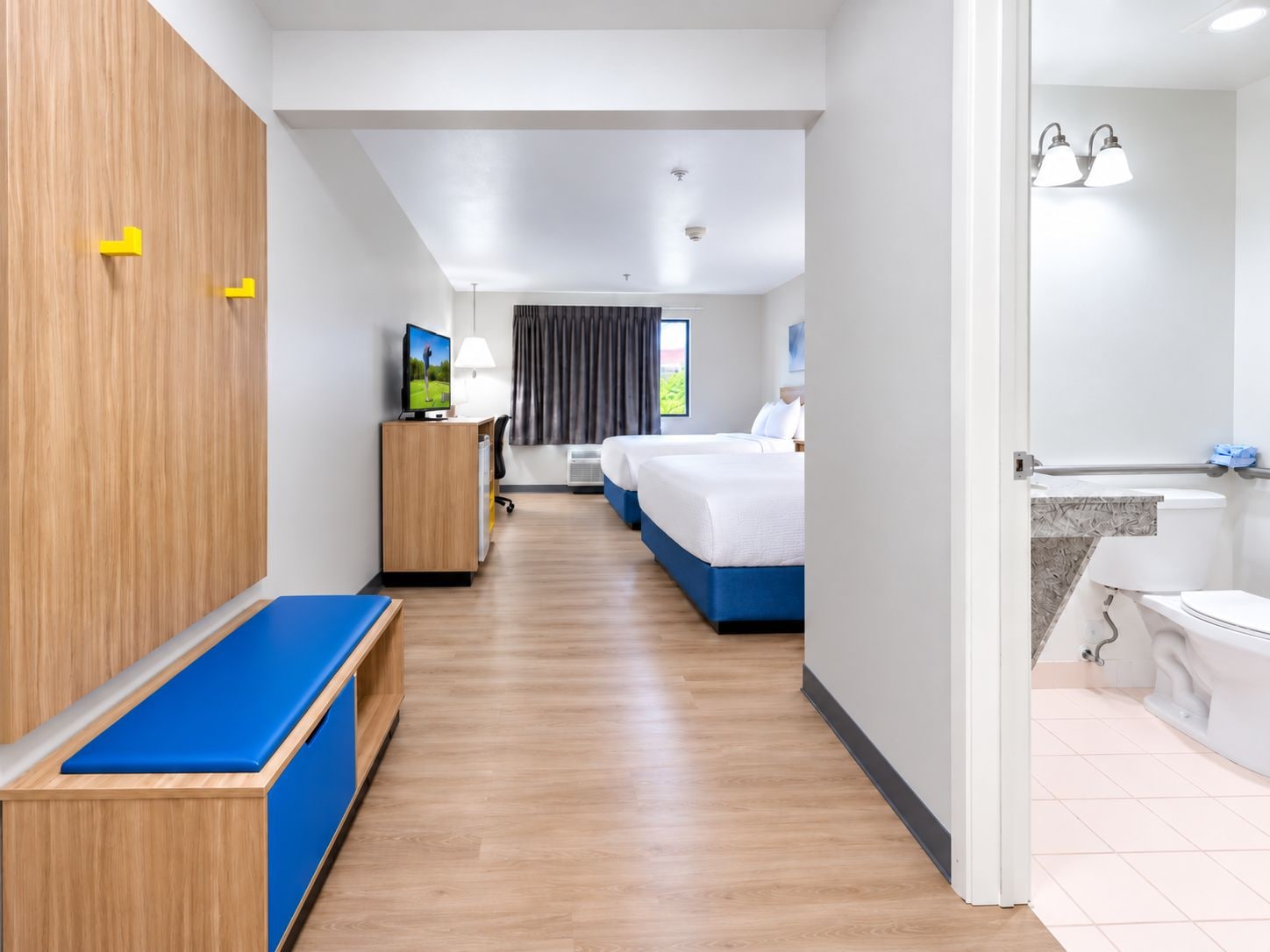 A hotel room with a wooden wardrobe, a desk with a television, a bed with a blue headboard, and a bathroom with a bathtub and toilet.