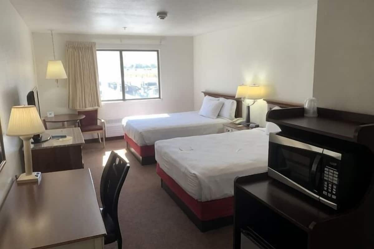 A hotel room with two beds, a desk, a chair, a microwave, and a window with curtains.