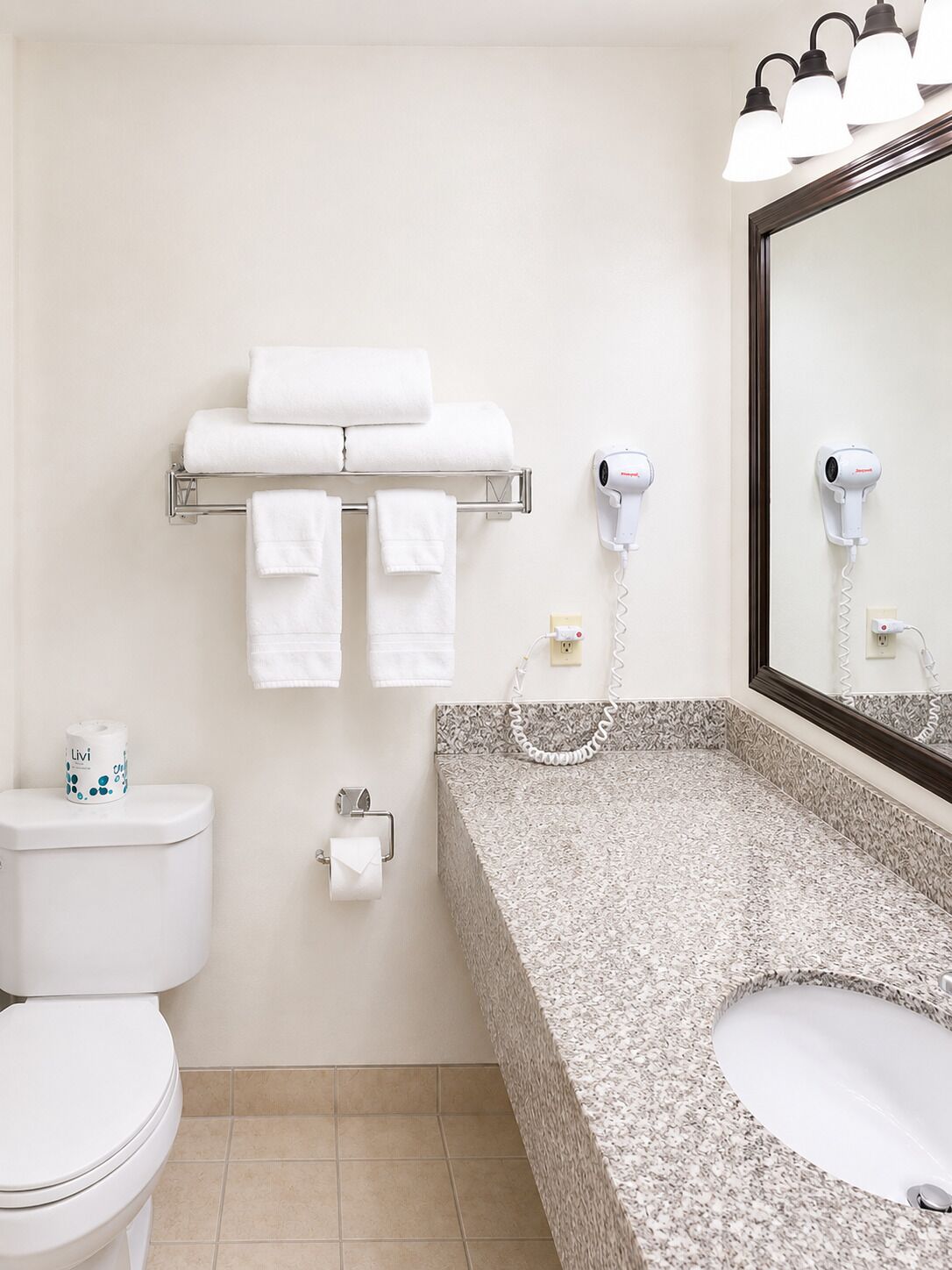 Un baño con inodoro, lavamanos con encimera de granito, toallero con toallas, secador de manos y espejo.