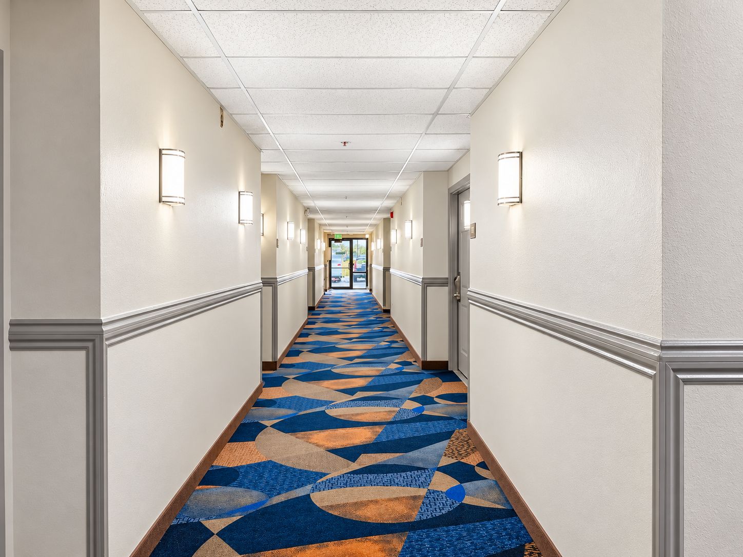 A hotel corridor with patterned carpet, wall-mounted lights, and doors on either side.