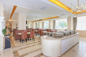 A spacious hotel dining area with a central buffet, multiple seating arrangements, and large windows providing natural light.
