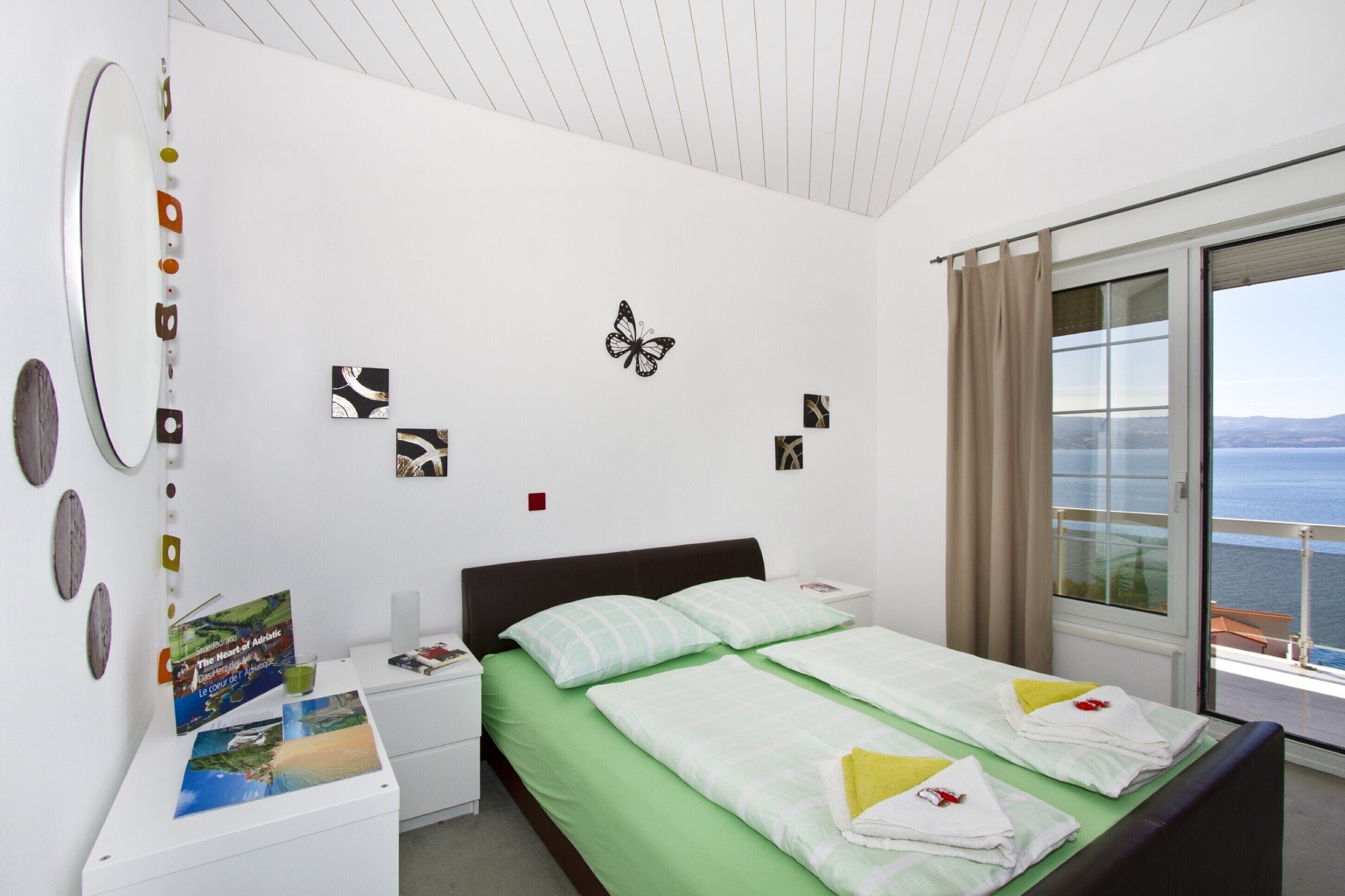 Une chambre d’hôtel comprenant un lit, une table de chevet, une fenêtre donnant sur la mer et des éléments de décoration murale, notamment un papillon et des tableaux encadrés.