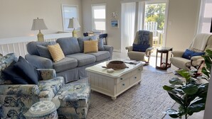 A living room with a grey sofa, a white coffee table, and a blue patterned ottoman. There are two lamps, a white chair, and a small table with a lamp. - 3 Bed/2.5 Bath 1450 sq ft with Large Open Floor Plan, 3 Min Walk to Beach (Emerald Isle)