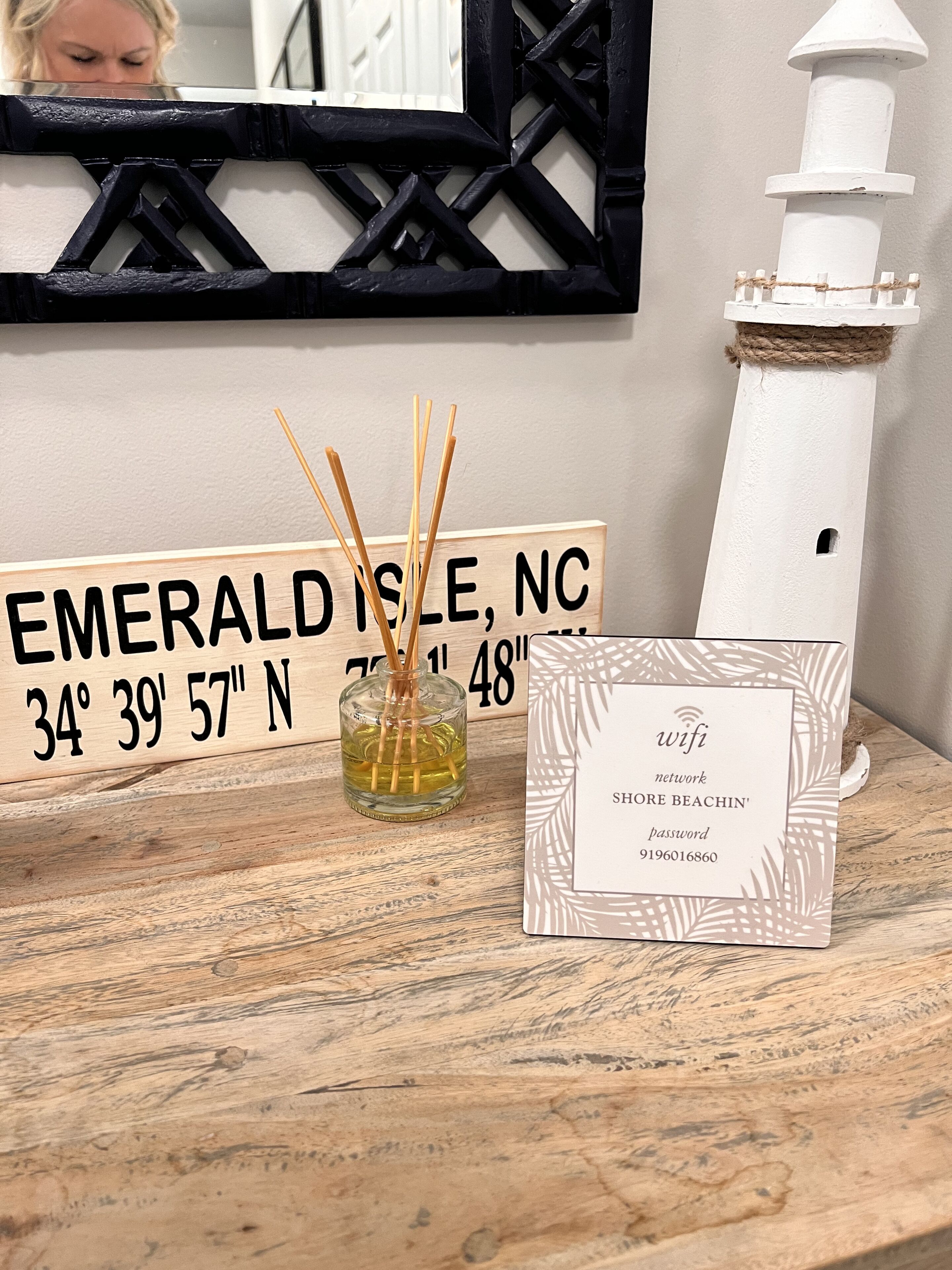 Een houten tafel met een bord waarop Emerald Isle, NC staat, een witte vuurtoren, een glazen vaas met rietstengels en een bord met het WiFi-wachtwoord.