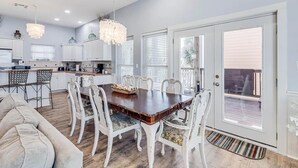 A spacious dining area with a large wooden table surrounded by white chairs, a kitchen in the background, and a sliding glass door leading to an outdoor deck.