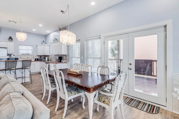 A spacious dining area with a large wooden table surrounded by white chairs, a kitchen in the background, and a sliding glass door leading to an outdoor deck.