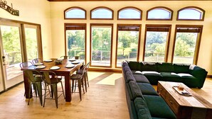 A dining area with a wooden table and chairs, a green sofa, and large windows offering a view of trees. - Waterfront Luxury Home on 5 Acres. 
Sleeps 22. Beds: 5 king, 4 queen, 4 twin XL (New Braunfels)