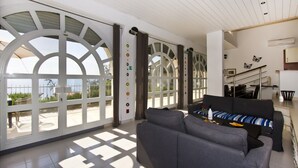 A spacious living area with a grey sofa, a dining table with chairs, and large windows with a view of the outdoors.