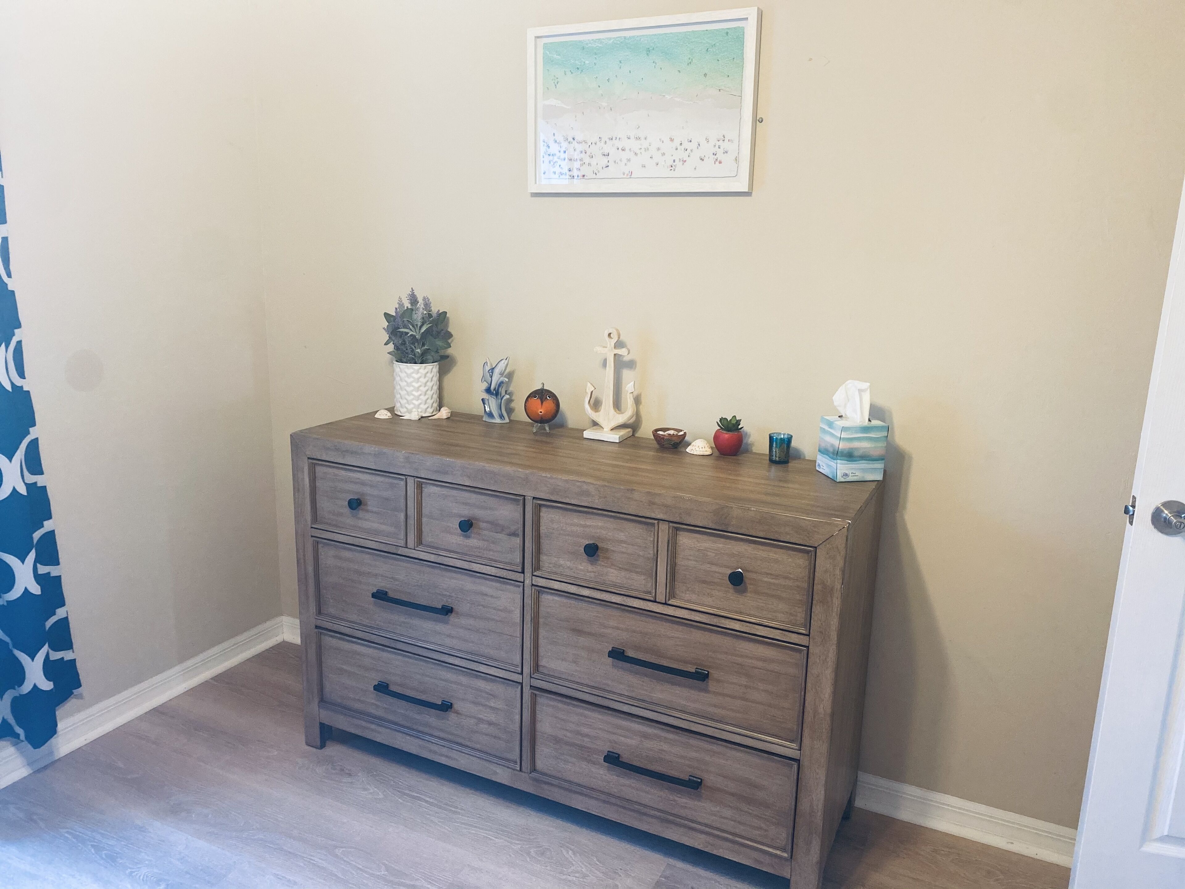 Un aparador de madera con seis cajones, una planta pequeña en maceta, objetos decorativos y un cuadro en la pared.