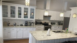 Uma cozinha moderna com armários brancos, uma ilha grande com banquetas, eletrodomésticos de aço inoxidável e iluminação pendente.
