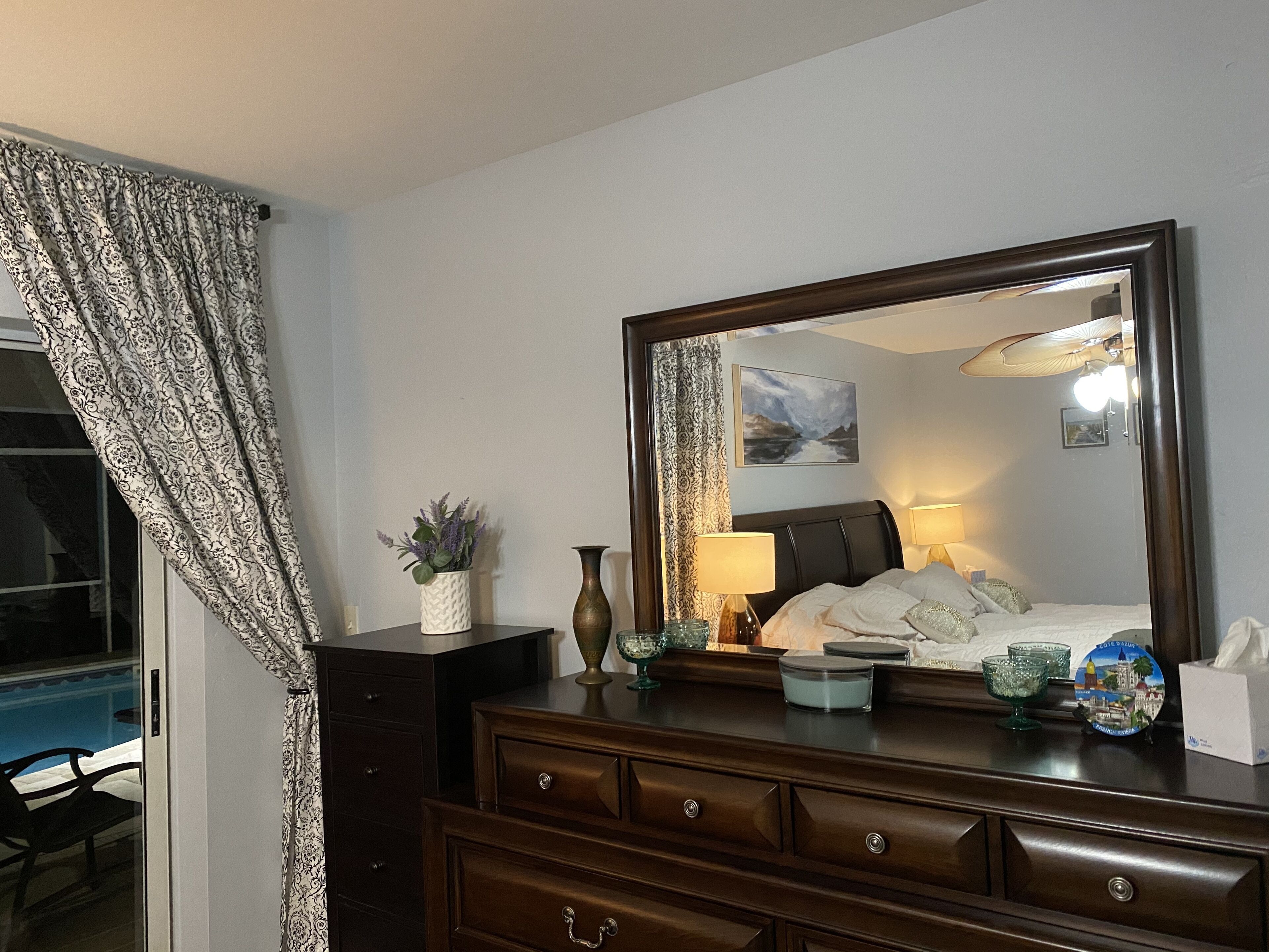 Habitación de hotel con un espejo grande, una cama con ropa blanca, un aparador con cajones, un jarrón con flores, una lámpara y una mesita con una vela y un vaso.