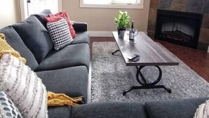 Ein Wohnzimmer mit grauem Sofa, einem Couchtisch, einem Kamin und einem Fenster mit Jalousien.