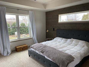 Une chambre à coucher avec un grand lit, une tête de lit en bois, une fenêtre avec des rideaux et une vue sur l’extérieur.