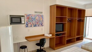 A compact room with a kitchenette, a wooden wardrobe, a desk with a chair, a television, and two beds. - Hotel Flat Alameda (Aracatuba)