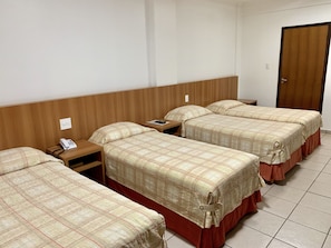 A hotel room with three beds, each with a plaid bedspread and wooden headboard. There is a bedside table with a phone and a remote control. - Hotel Flat Alameda (Aracatuba)
