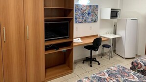 A bedroom with a wooden wardrobe, a bed with patterned bedding, a desk with a chair, a television, a microwave, and a refrigerator. - Hotel Flat Alameda (Aracatuba)