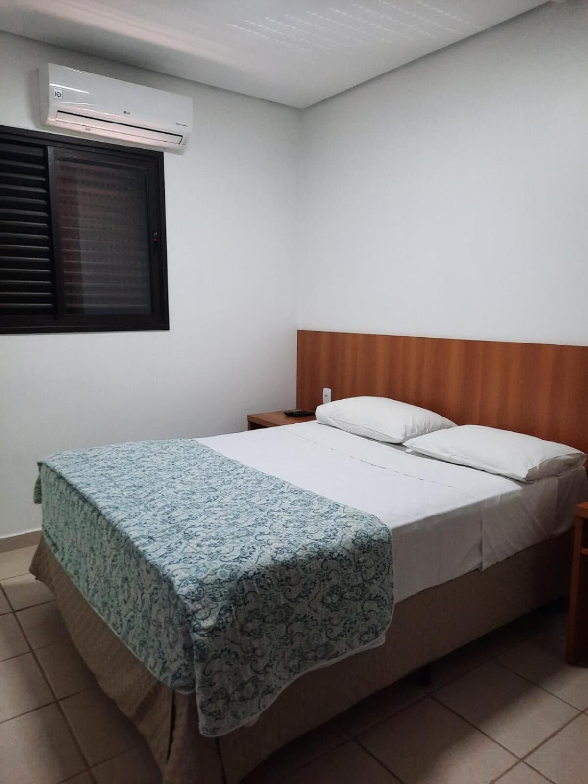 Un dormitorio con una cama, una mesita de noche, un aire acondicionado y una ventana con persianas.