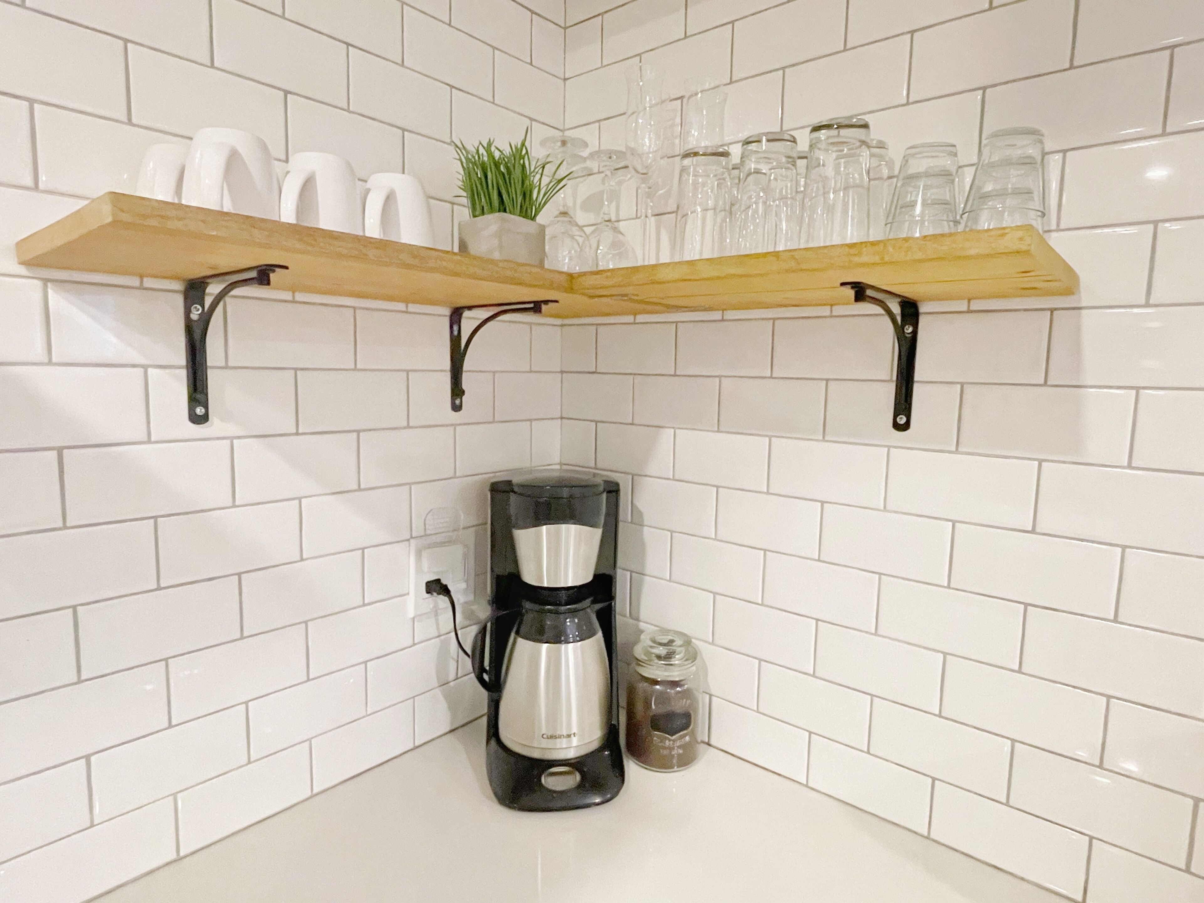 Uma cozinha com paredes revestidas de azulejos brancos, duas prateleiras de madeira com copos, uma cafeteira e um frasco de temperos.