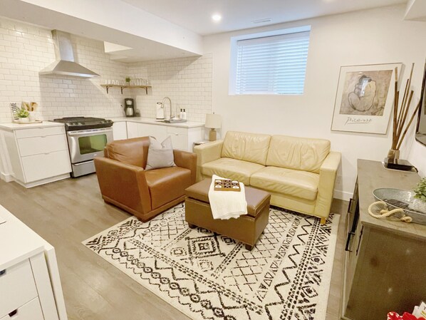 Un espace de vie compact comprenant un coin cuisine, un canapé en cuir marron, un canapé beige et un tapis à motifs.