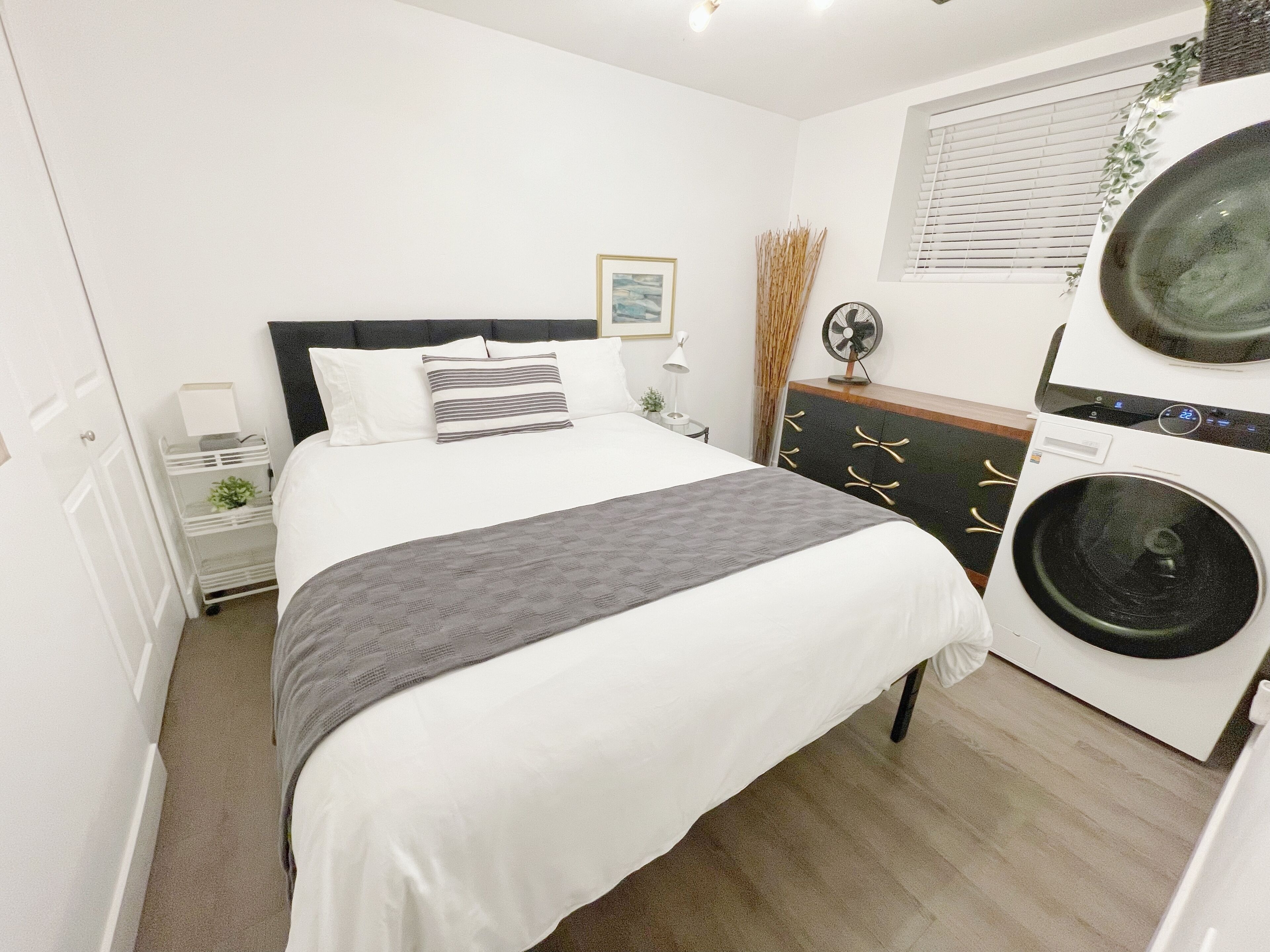 A bedroom with a bed, a bedside table, a dresser, a fan, and a washing machine.