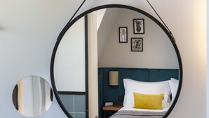 Un dormitorio con un espejo circular colgante, una cama con almohadas blancas y amarillas, un cabecero color aguamarina y cuadros en la pared.