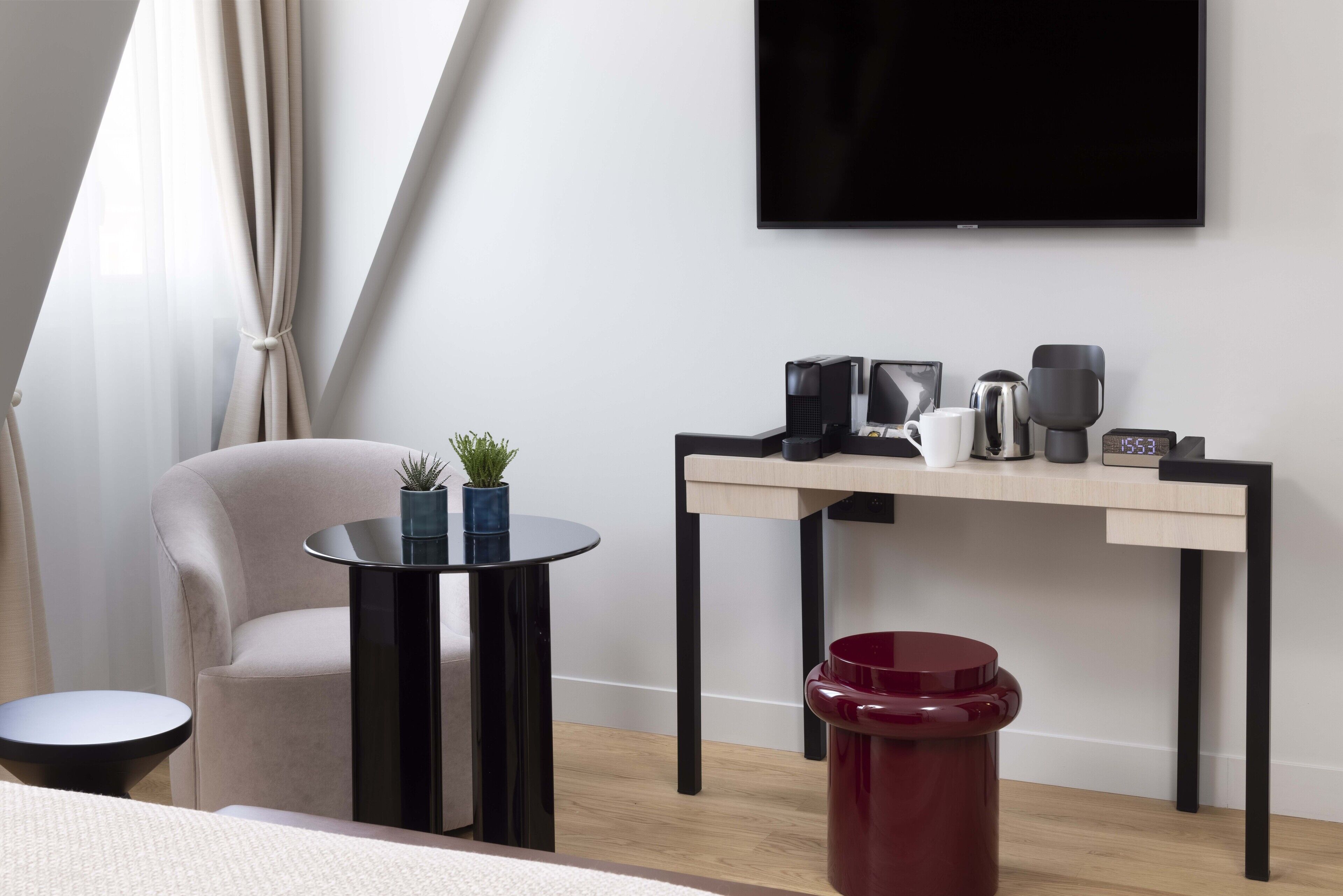A modern hotel room with a flat-screen TV mounted on the wall, a small round table with a potted plant, a red trash bin, and a wooden desk with a coffee maker and a clock.