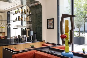A modern bar area with shelves of bottles and glasses, a coffee machine, and seating with orange cushions.
