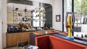 Un área de bar moderna con estantes llenos de botellas y vasos, una zona de estar con cojines naranjas, y un ventanal con vistas a árboles.