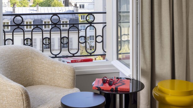 Una habitación con vistas a edificios a través de una ventana, un sillón beige, una mesita negra con un libro rojo y auriculares, y un cubo de basura amarillo.