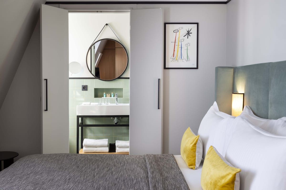 A modern hotel room with a bed, a bedside table, a mirror, a bathroom visible through an open door, and a framed artwork on the wall.