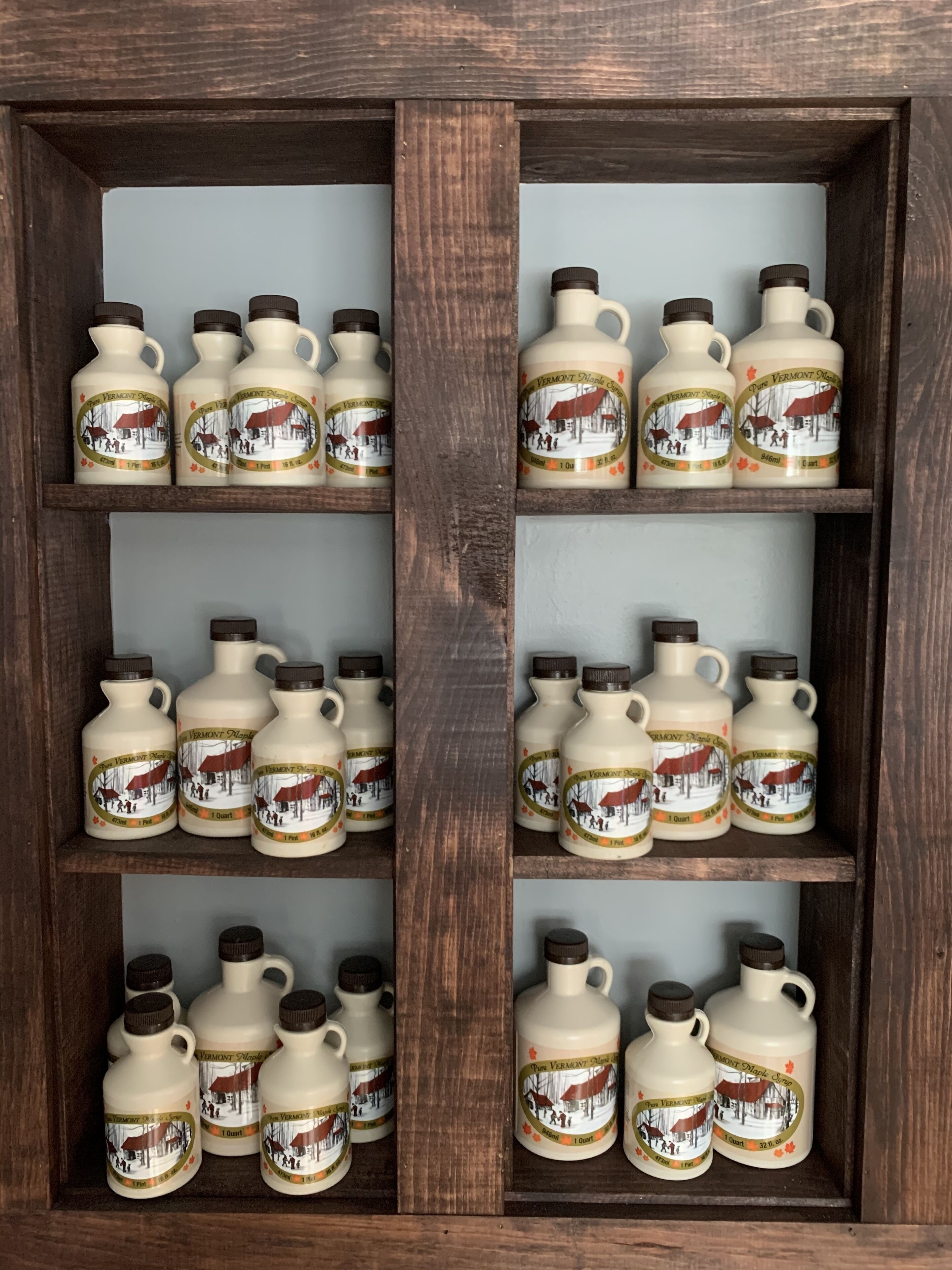 A wooden display case with multiple shelves, each containing white ceramic bottles with brown caps and labels depicting a snowy village scene.