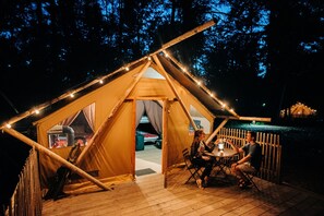 Une tente dotée d’une terrasse en bois, de guirlandes lumineuses et d’une table où sont assis deux personnes.