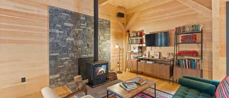 A cozy living room with a stone wall, a fireplace, wooden furniture, and a bookshelf.