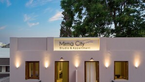 Un edificio con un cartello che riporta la scritta "Mirma City Studio & Apartment". Sono visibili quattro finestre con luci, un balcone con sedie e una vista su alberi e un edificio sullo sfondo.