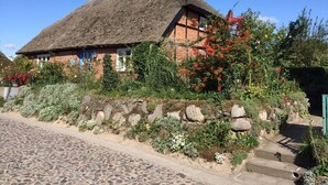 A traditional thatched-roof house with a stone wall and colorful flowering plants. - Vacation home Südstrand **** Use of swimming pool, exclusive house with own sauna (Göhren)