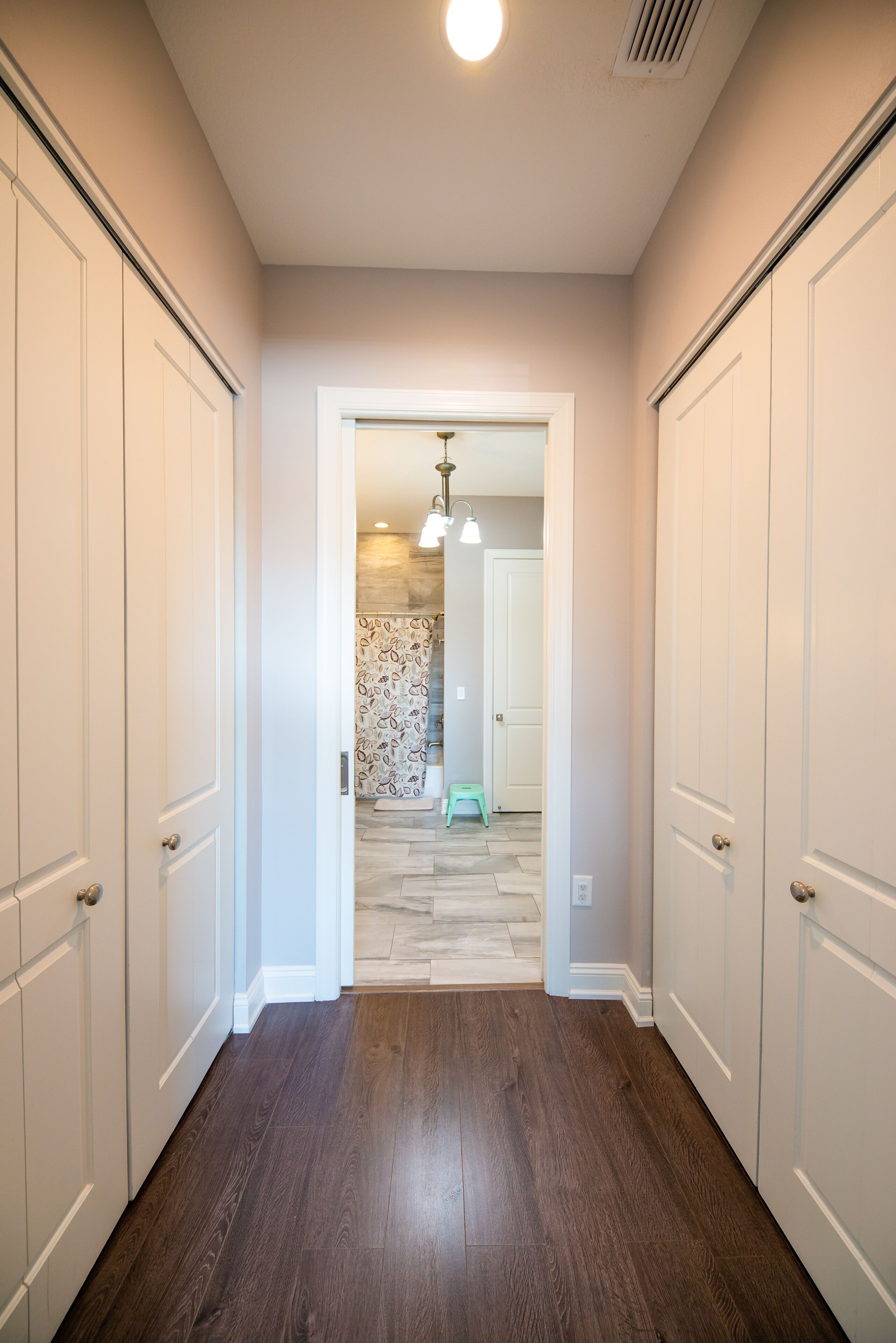 A hallway with wooden flooring, white doors, and a view into a room with patterned wallpaper and a hanging light fixture.