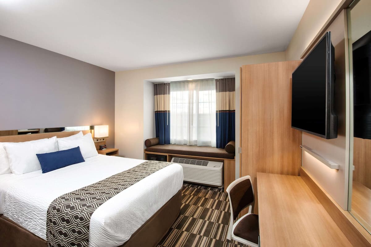A hotel room with a large bed, a desk, a chair, a flat-screen TV, a window with curtains, and a wall-mounted TV.