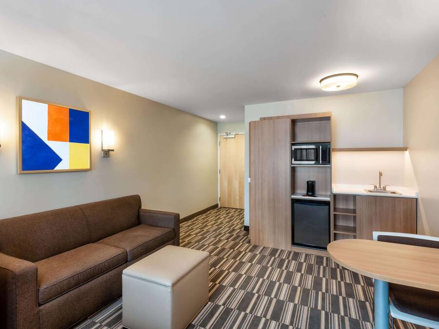 A compact hotel room with a sofa, a small table, a kitchenette, and a wall-mounted painting.